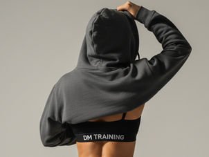 Een vrouw die met één hand een oversized hoodie over haar hoofd trekt of net uittrekt, terwijl daaronder een DM-outfit zichtbaar wordt.