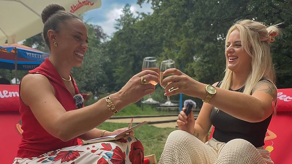 Twee vrouwen lachen en klinken met een glaasje rosé tijdens een openluchtinterviewmoment — een sfeerbeeld van warme herkenning en verbinding.