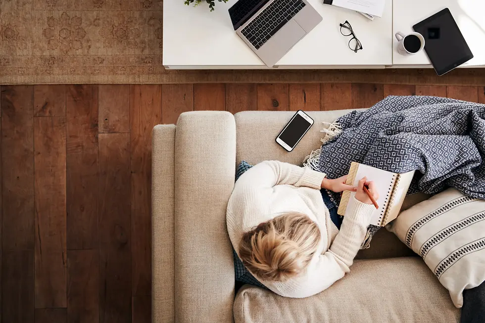 foto panorâmica de uma mulher branca, loira, influencer digital sentada no sofá de sua sala escrevendo num caderno; ao seu lado, no sofá, um iPhone e numa mesa de centro, o seu notebook e um óculos; a proposta dessa foto é mostrar que os influenciadores digitais podem trabalhar de qualquer lugar, te conforto e liberdade geográfica.