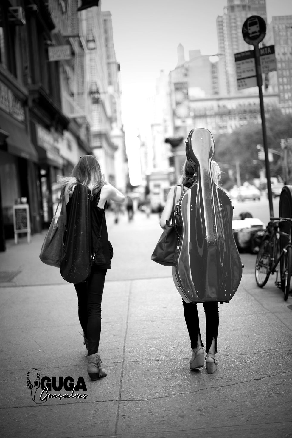 duas mulheres de costas andando com as bags de seus respectivos instrumentos nas costas, uma com um violino e a outra com um violoncelo. A foto tem o objetivo de ilustrar duas musicistas a caminho de tirar a OMB.