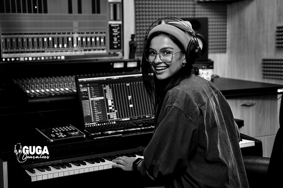 foto de uma jovem mulher sorridente, de costas, olhando para trás, dentro de um home-studio de produção musical, onde mixa uma trilha sonora; no estúdio, uma mesa de som, interface, monitor de vídeo grande, interface de áudio e demais aparatos.