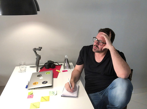 Foto de Guga Gonçalves, sentado a frente de sua mesa, escrevendo anotações num caderno; sobre a mesa, notebook fechado, tripé, câmerca fotográfica Canon, pasta e postites; Guga está com a mão na cabeça com um ar reflixivo; a imagem tem o objetivo de conectar o leitor com a ideação da Mentoria Personal Branding.