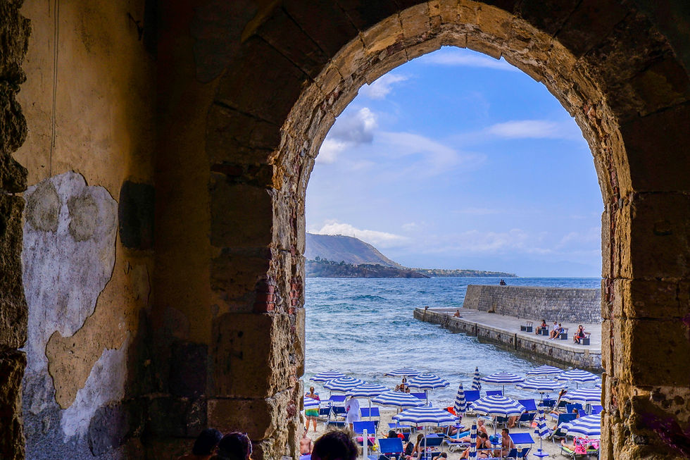 Stone arch frames a beach with blue-striped umbrellas and people relaxing. Ocean and mountain visible in the background under a clear sky.