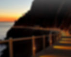 Cliffside path at sunset with glowing lights along the railing. Dark silhouette of a mountain and calm ocean in the background. Peaceful mood.
