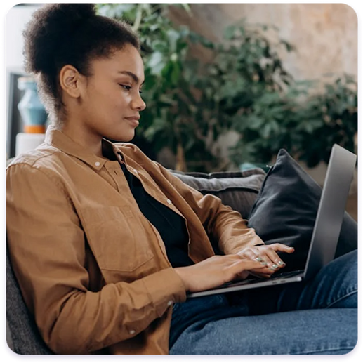 Woman with hair in a bun wearing a brown shirt, sitting comfortably on a couch and working on a silver laptop.