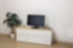 A minimalist room features a white cabinet with a clock, a computer monitor, and magazines on top, next to a potted plant on a wooden floor.