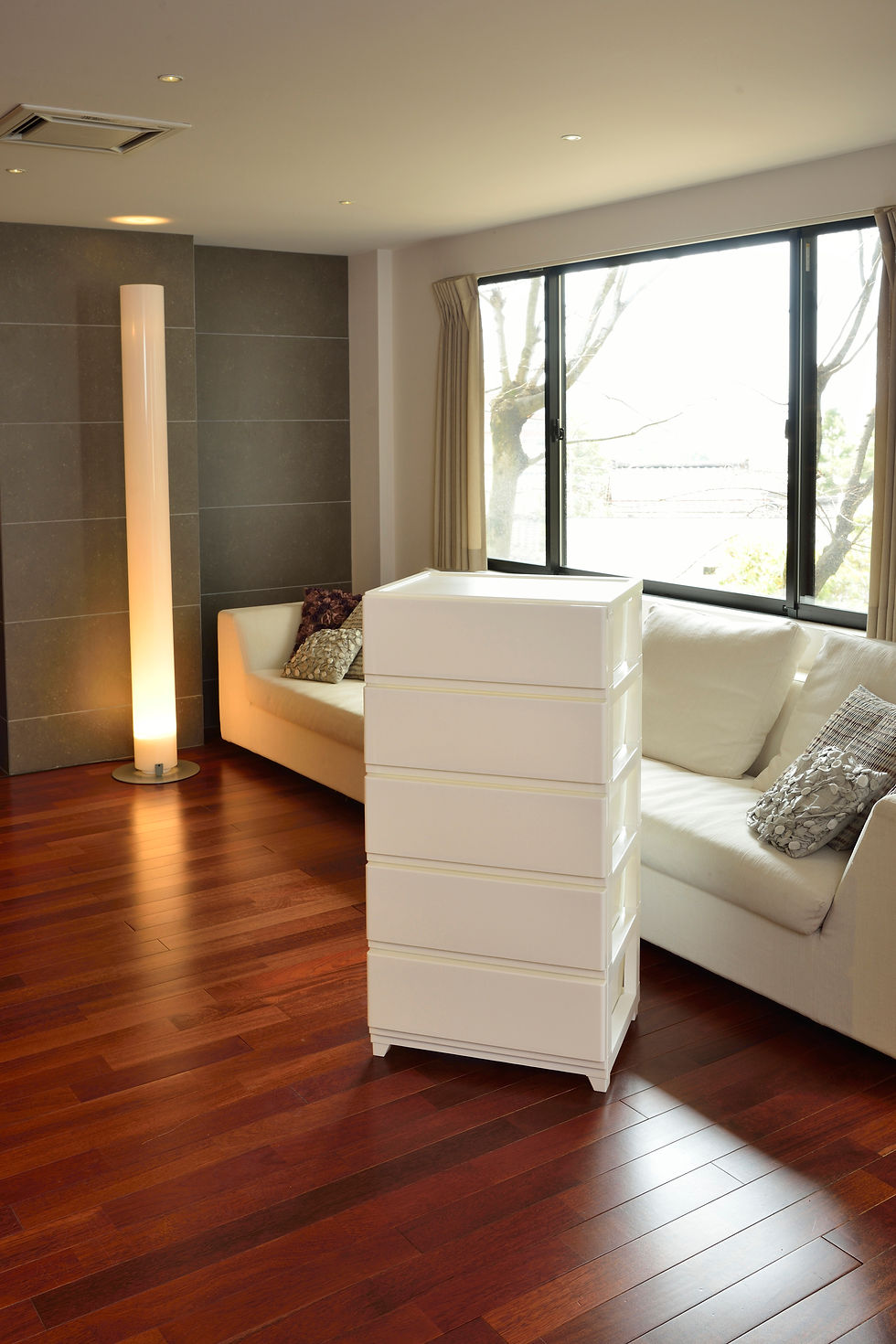 White drawers in a modern living room with a white sofa, cushions, tall lamp, wooden floor, and large window showing trees outside.