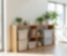 Wooden shelf with potted plants, beige storage bins, and books in bright room with large windows and light wood flooring. Minimalist decor.