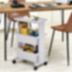 White rolling cart with books and stationery beside a wooden desk. Camera and notebooks on the desk, leather chairs. Bright, organized space.
