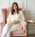 A woman wearing ivory shirt and white pants sitting in a velvet pink chair looking directly at you.