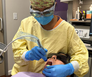 Dentist in yellow gown and patterned cap examines a patient's teeth using dental tools. The patient wears sunglasses. Medical setting background.