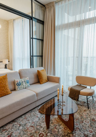 Living room from the dining side — sofa, biomorphic coffee table, bouclé chair, balcony, and bedroom visible through the glass partition