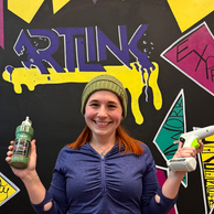 Smiling woman holds paint bottle, glue gun near ART INK wall.