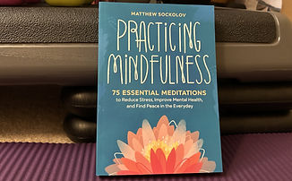 A copy of 'Practicing Mindfulness' by Mathew Sockolov resting on a purple yoga map with a step up and multicolored hand weights in the background
