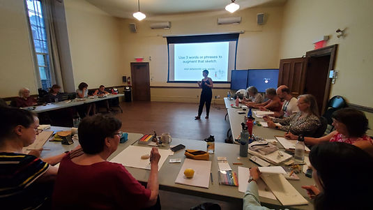 A person stands in the center of a room, in front of a large screen upon which a presentation is being projected. The presenter is speaking to a room of people seated at a set of tables organized in a "u" shape. People at the tables are drawing nature objects like leaves, bones, and lemons.