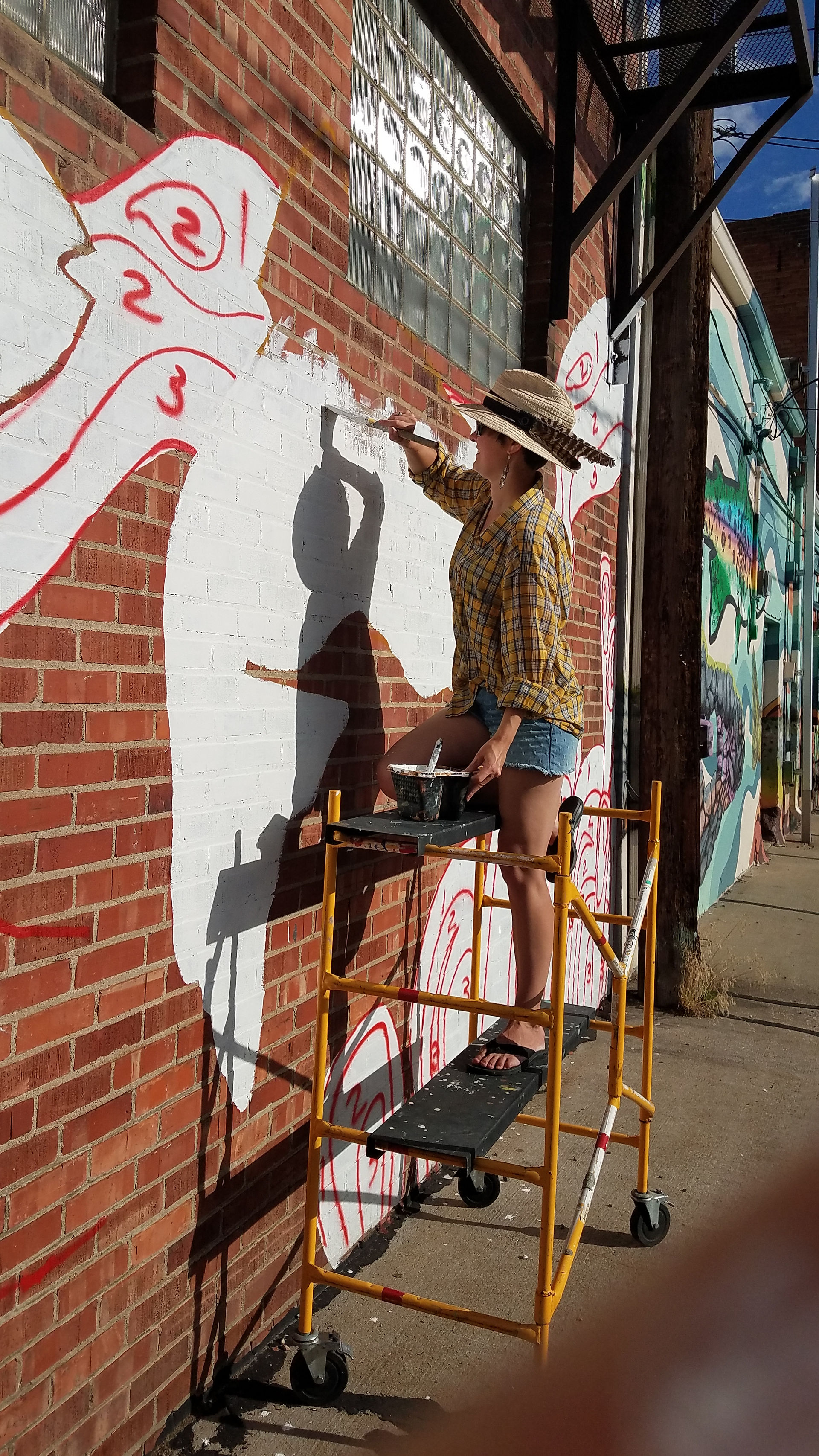 Woman balances on scaffolding in order to paint part of a mural on a wall