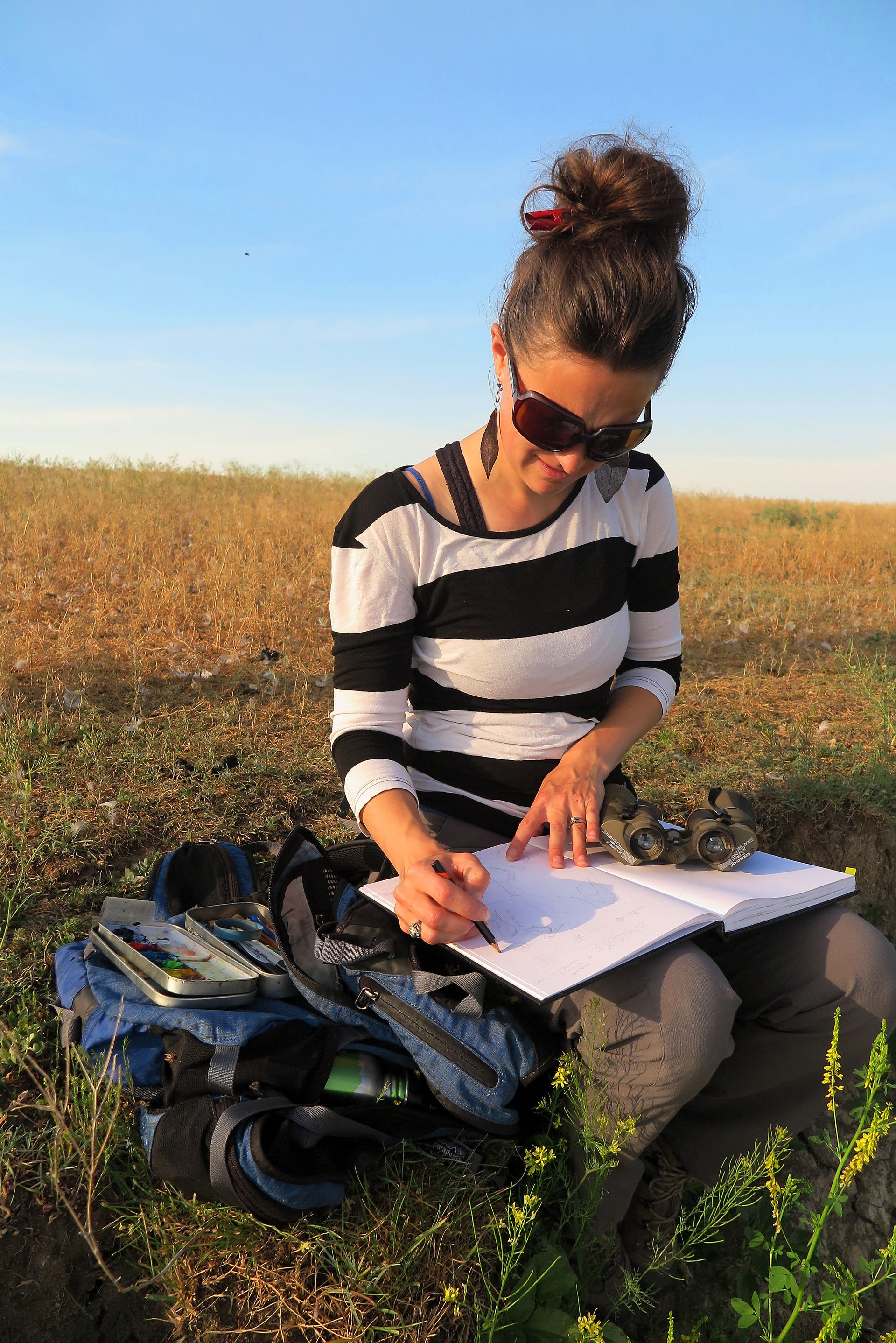 Woman sits in a field, looking down at the sketchbook she is drawing in