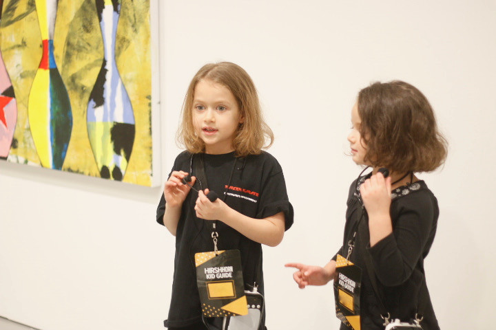 Two young girls carrying portable microphones stand next to an artwork and talk about it to a group of people. The group is out of the frame.