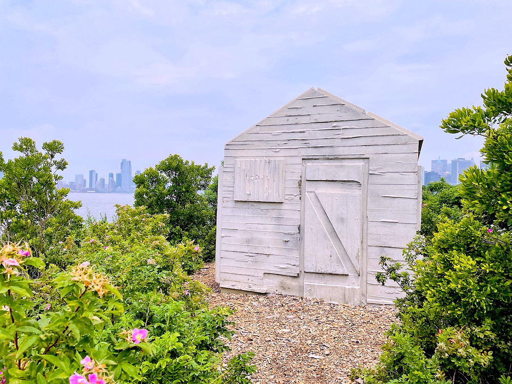With the backdrop of the New York City Skyline, a small, white, wooden, house-like structure called the Cabin, a permanent installation by British artist Rachel Whiteread, is surrounded by plants and pink flowers.