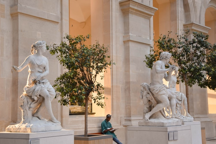 A person, surrounded by marble statues, sitting inside a museum listening to a museum audio guide with headphones on.