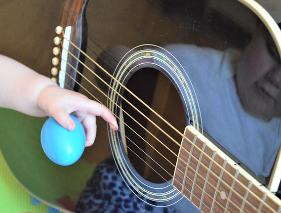 Image of the body of a black guitar on its side, there is the reflection of a baby looking at the guitar on the instrument. The hand of the baby is holding an egg shaker and has a finger on a guitar string.