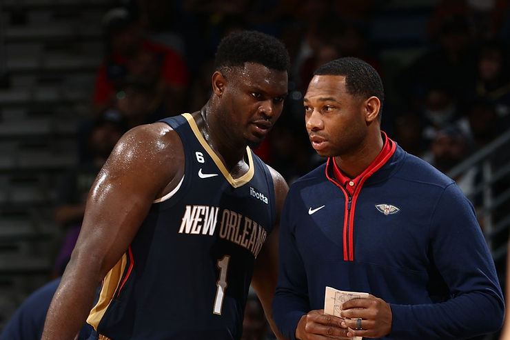 Zion Williamson talking with a coach during a basketball game, both wearing New Orleans Pelicans uniforms.