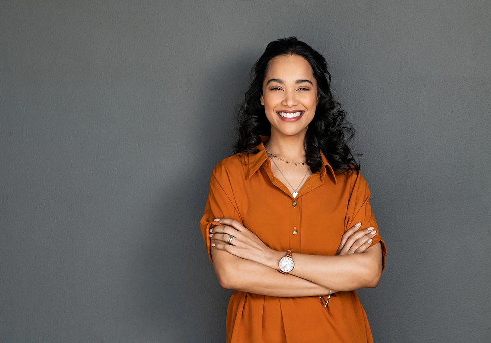Smiling coach with crossed arms in an orange shirt, standing against a gray wall. She exudes confidence and wears a watch and necklace.