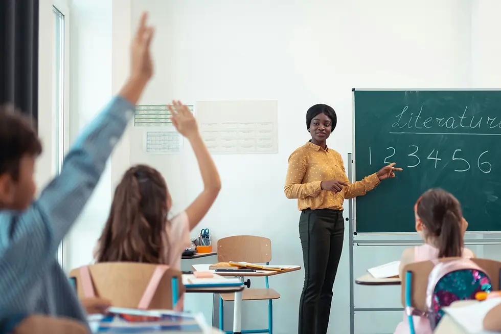 Female teacher standing in front of elementary class point at green chalk board while looking at students raising their hands