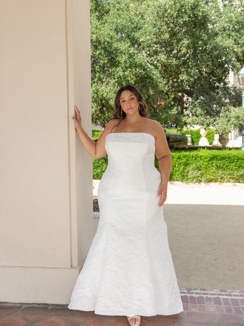 Plus size bride wearing a strapless fit and flare wedding gown with a textured fabric design, standing outdoors against a column.