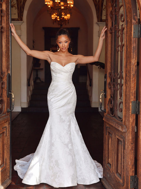Bride wearing a fit-and-flare satin wedding dress with a sweetheart neckline, ruched bodice, and elegant chapel-length train, standing in double wooden doors.