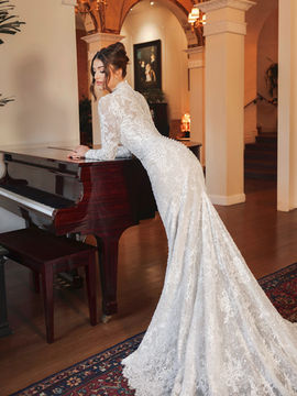 Bride in a form-fitting long-sleeve lace wedding dress featuring a high neckline, intricate lace detailing, and a dramatic train, posing by a grand piano indoors.