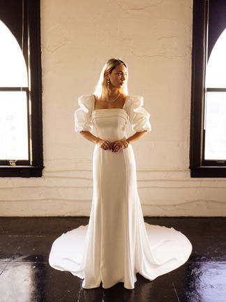Bride in a modern wedding gown featuring puff sleeves, a square neckline, and sleek silhouette with flowing train, standing between tall windows.