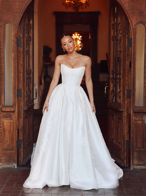 Bride in a strapless ball gown wedding dress standing in a doorway, featuring a fitted bodice and a voluminous skirt with elegant pleats.