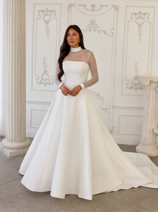 Bride in a high-neck wedding gown featuring sheer long sleeves, a fitted bodice, and a flowing A-line skirt with train, posing in a classical interior with white columns and ornate walls.