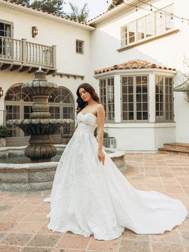 Bride wearing a strapless jacquard ball gown wedding dress featuring a fitted bodice, full skirt, and elegant long train, standing outdoors near a fountain.