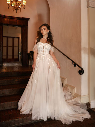 Bride in an elegant off-shoulder tulle wedding gown featuring a structured corset bodice with floral appliqués and a flowing train, posing gracefully near a staircase.