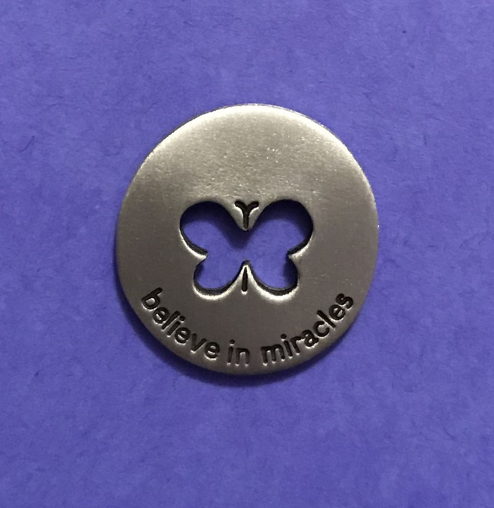 A circular metal coin with a butterfly cutout and it reads Believe in Miracles set on a pale green background.