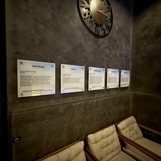 Lounge seating area at Be Well Studio with wall-mounted treatment descriptions for hot tub, cold plunge, and other wellness therapies beneath a large decorative clock.