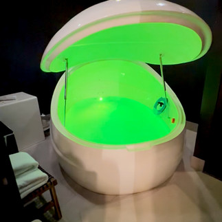 Float pod filled with glowing green water, partially open in a private room with a towel rack and minimal decor for sensory isolation sessions.