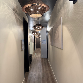 Bright hallway inside Be Well Studio featuring modern woven pendant lighting, light wood floors, and clean beige walls lined with treatment signage.