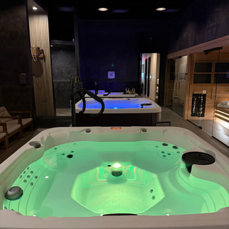 Dimly lit hydrotherapy zone at Be Well Studio featuring two glowing hot tubs and adjacent sauna cabins, offering a serene recovery atmosphere.