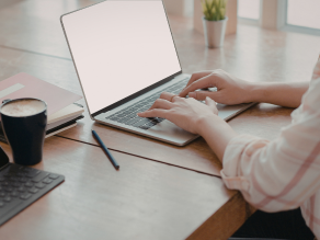 The image depicts a person sitting at a desk, working on a laptop. The individual is using both hands to type on the keyboard. On the desk, there are several items including another laptop, a cup of coffee, a pink notebook, and a pen. The setting appears.