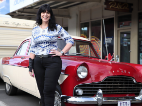 Justine Martin smiling and standing confidently in front of a vintage red and cream car parked on a street, wearing a patterned top and black pants, with a wrist brace on her right hand.