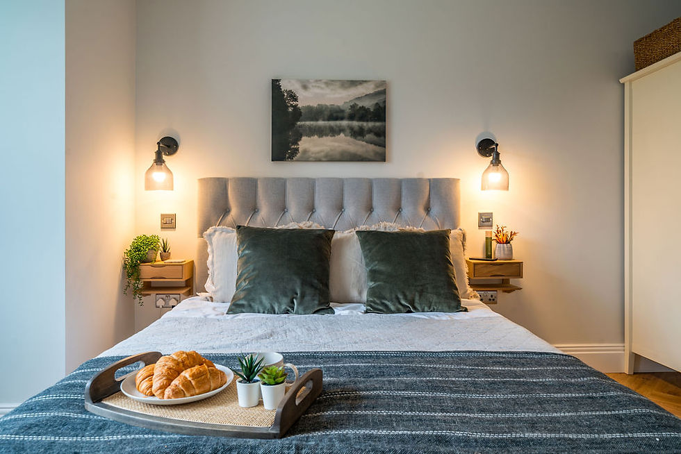 Cozy bedroom with a plush bed, green cushions, and a tray of croissants. Warm lighting, a photo on the wall, and a tranquil atmosphere.