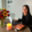 Photo of writer nuris sitting with her hand on her laptop and one under her chin smiling at the camera. Flowers, a candle, and books surround the computer