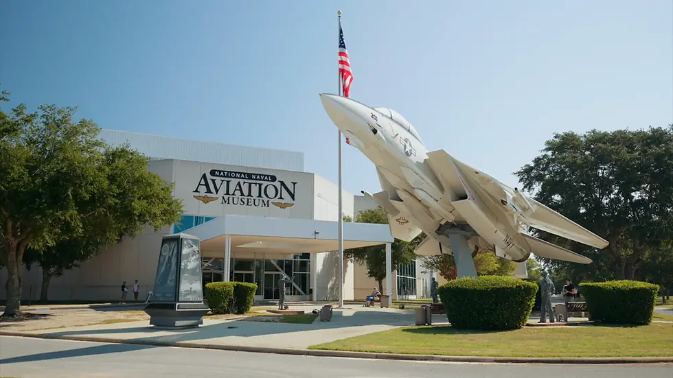 Jet displayed outside National Naval Aviation Museum. U.S. flag and statues in foreground. Building with large sign, clear sky, trees nearby.