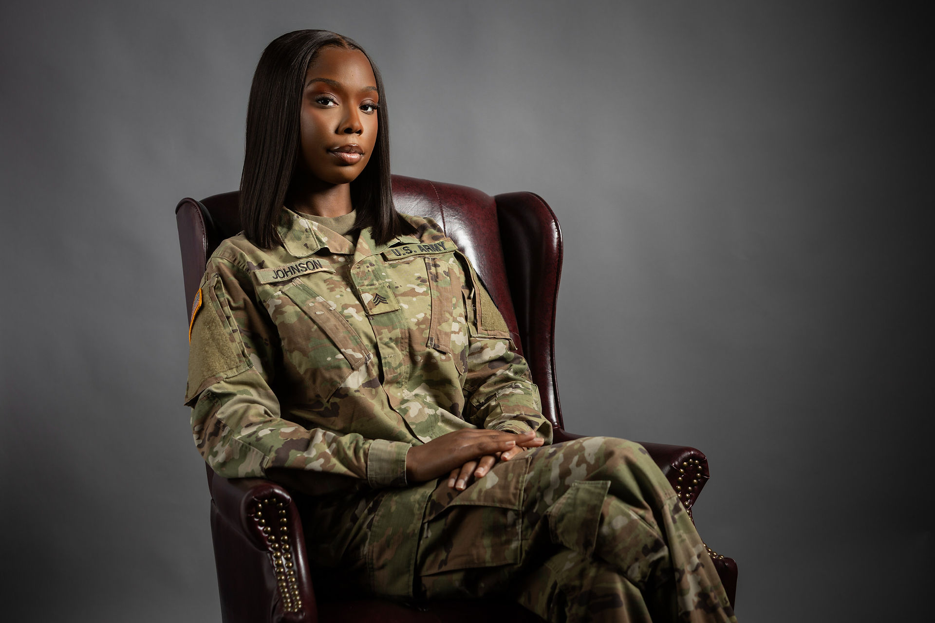 Female Army soldier in OCP camouflage uniform seated in leather wingback chair, cinematic studio portrait by Austen Hunter Photography.