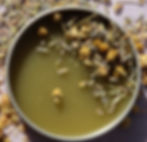 Round tin containing a green salve on a lavender cloth surrounded by chamomile flowers