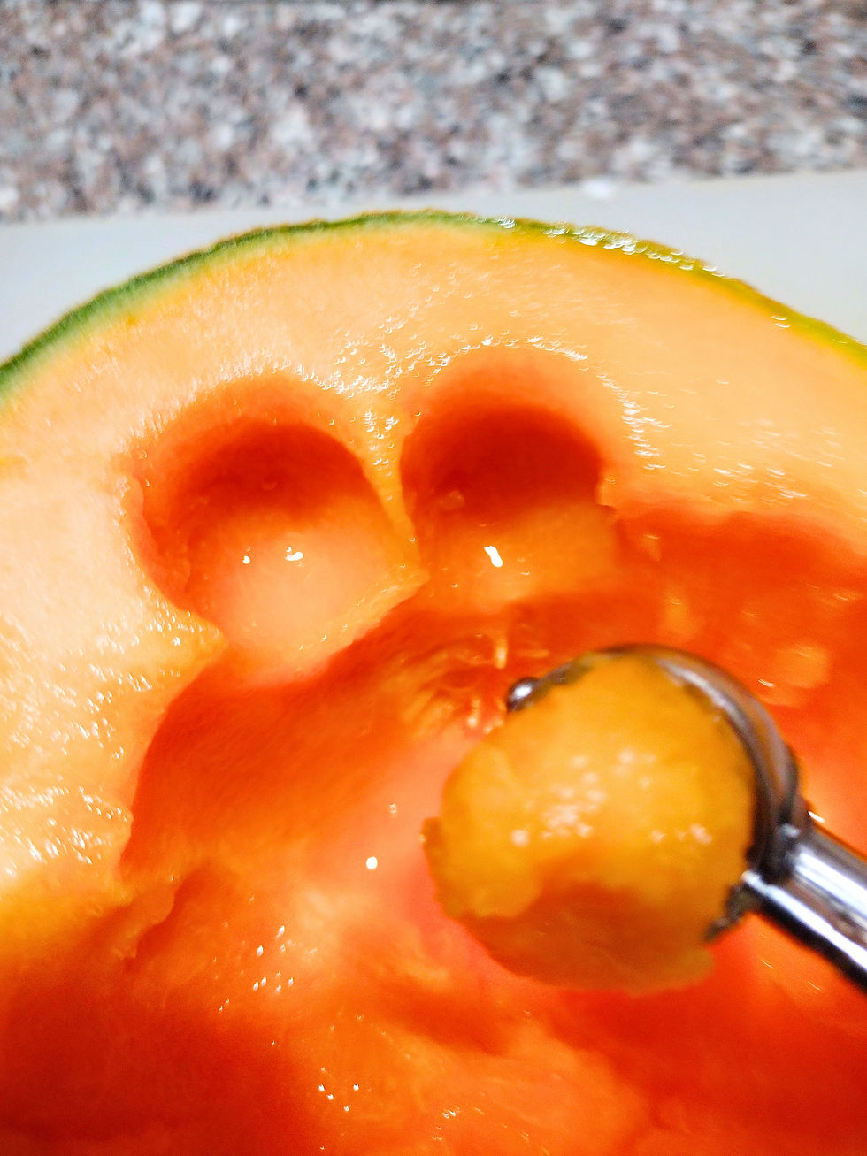 Close-up of a cantaloupe with melon baller creating round scoops. Orange fruit flesh and rough brown counter in the background.