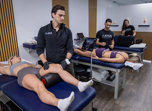 Therapists treat two women on massage tables in a clinic. "AVION" logo visible. Using Diagnostic Ultrasound and cupping therapy. Modern setting with wood paneling and equipment.